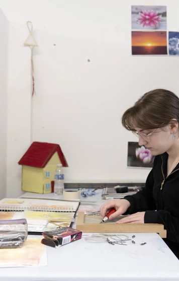 Person seated at a desk in an art studio, carving a design into a printmaking block, with sketchbooks and artwork on the wall Person seated at a desk in an art studio, carving a design into a printmaking block, with sketchbooks and artwork on the wall